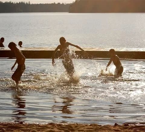 En grupp människor njuter av en solig dag vid sjön på Jälluntofta camping. Några av dem är barfota och tar del av olika fritidsaktiviteter på stranden.