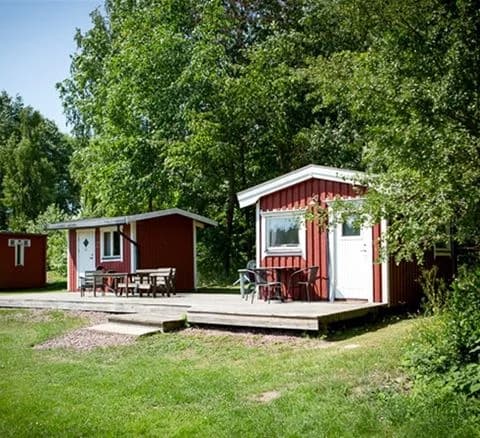 Bild av Ivö Camping: En grönskande campingplats med stora gräsytor, omgiven av natur i vårens skrud. Flera små stugor står utspridda i området med träd och buskar som ger skugga. Området har en trädgårdslik känsla med inslag av parkmiljö.