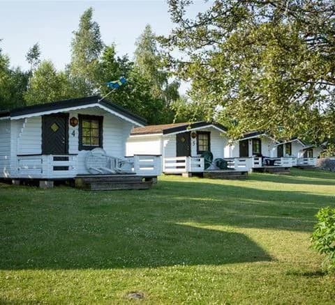En mysig stuga omgiven av en grönskande gräsmatta och trädgård på Ivö Camping, med skuggande träd och en lantlig atmosfär.