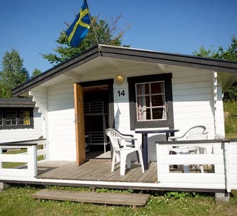 En trästuga med en veranda på Ivö camping, omgiven av trädgård och utemöbler.