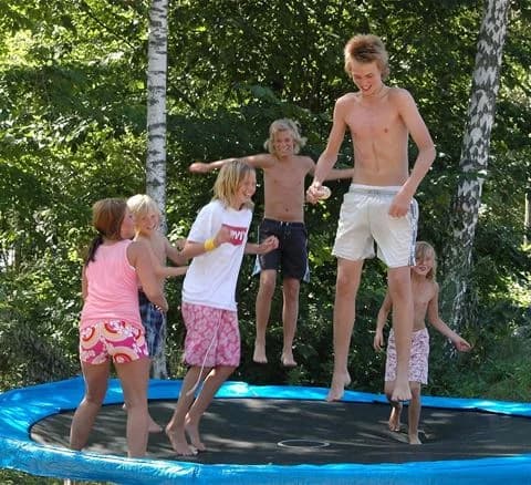 Barn och vuxna hoppar barfota på en stor studsmatta i soligt sommarväder, omgivna av grönska vid Ivö Camping.
