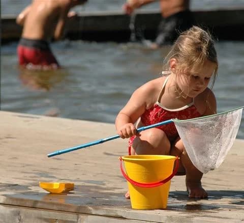 Ett litet barn i sommarkläder leker barfota med en hink utomhus på Ivö camping. Barnet verkar njuta av solen och omgivningen under en avslappnad semester eller rekreation.