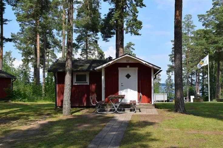 En pittoresk stuga omgiven av träd och välskött gräsmatta på Hätte camping, med små trädgårdsbyggnader i bakgrunden.