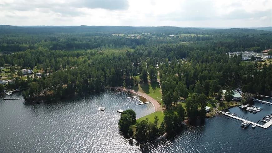 Flygbild av Hätte Camping vid en vattenväg, där en småbåtshamn med bryggor och båtar syns. Omgivande kustlandskap och inlopp ger en naturskön vy.