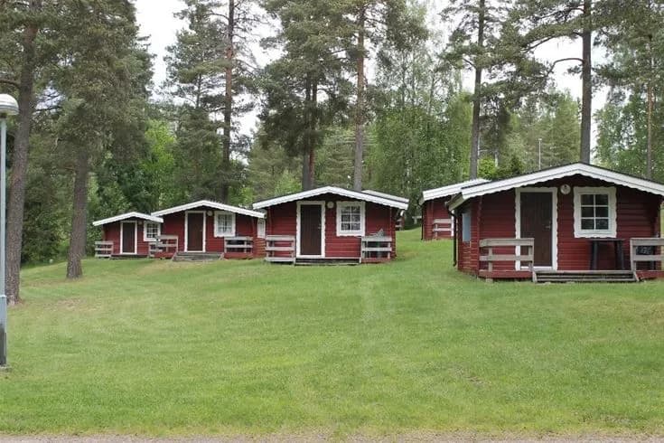 Bild av Hjortsjöns camping, visar en idyllisk campingplats med grönt gräs, pittoreska timmerstugor och trädgårdsbyggnader omgivna av lummiga träd och skuggiga ytor.