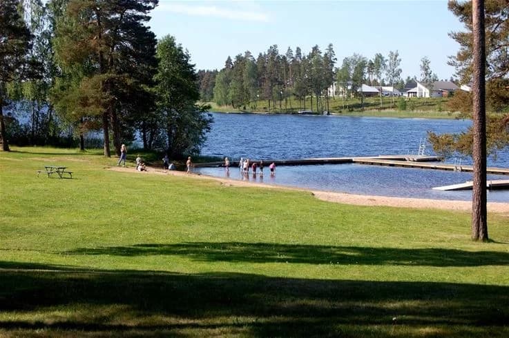 Bild av Hjortsjöns camping som visar en idyllisk sjö med klart vatten omgiven av gröna träd. Strandkanten bjuder in till rekreation och avkoppling i naturen.