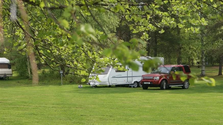 Alt-text: En campingplats vid Hedesunda med flera husvagnar och campervans parkerade på grön gräsyta. Området omges av träd vilket ger en naturskön atmosfär.
