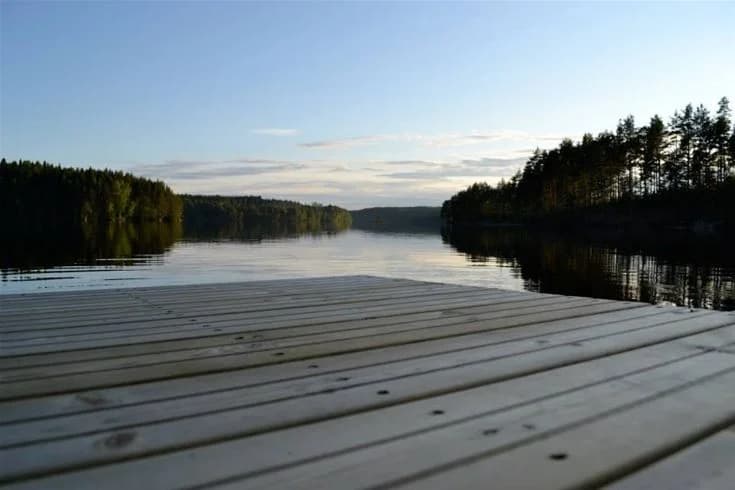 En träbrygga sträcker sig ut över en stilla sjö omgiven av grönska, medan moln flyter på en klar himmel ovanför.
