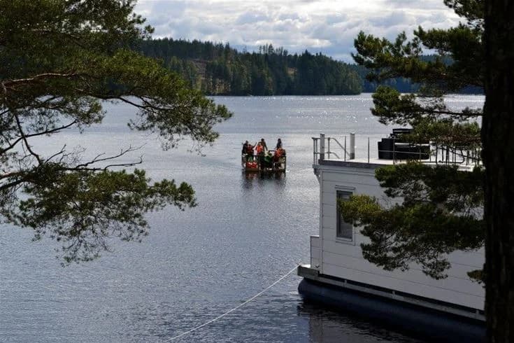 En lugn sjö vid Grums Fish n' Camp - Sävsjön, omgiven av grönskande träd. En båt ligger förtöjd vid stranden, redo för äventyr på vattnet.