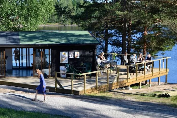 En mysig stuga vid Sävsjöns strandkant, omgiven av frodig grönska med en brygga som sträcker sig ut över vattnet. Utemöbler står på bryggan, redo för avkoppling med utsikt över den natursköna sjön.