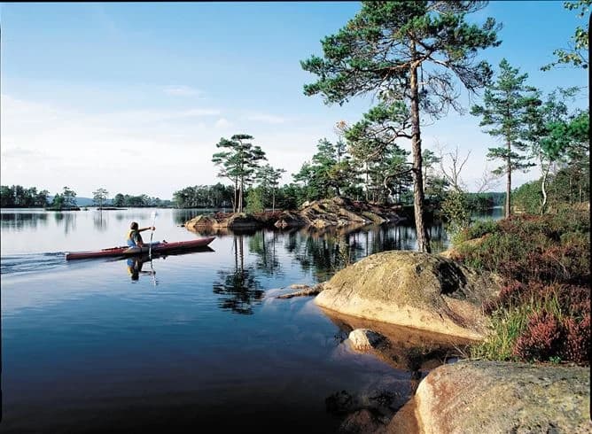 En naturskön sjö med spegelblankt vatten, omgiven av frodig gröna skogar. Två kanoter flyter stilla nära strandkanten vid Glaskogens vildmarkscamping.