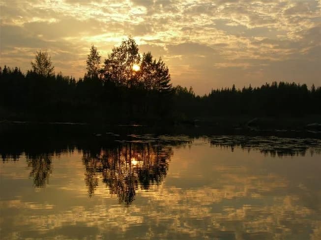 En lugn sjö vid solnedgången omgiven av tät skog i Glaskogens vildmarkscamping, med himlen färgad i varma toner och speglingarna av träden och himlen i vattnet.