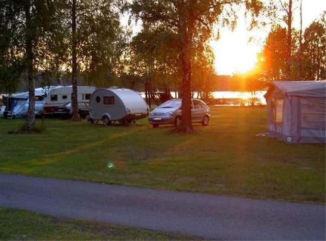 En husbil står parkerad vid e4:ans camping i skymningen, omgiven av tält och träd. Solnedgångens ljus reflekteras i en närliggande vattenyta, vilket skapar en lugn och fridfull atmosfär på campingen.