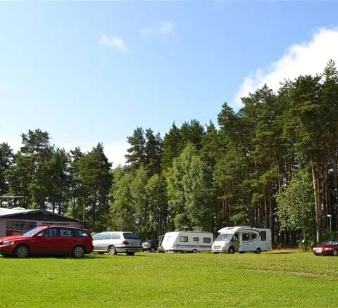 En parkering vid Djupadalsbadet med flera bilar, inklusive familjebilar och en husbil, omgiven av träd och med registreringsskyltar synliga.