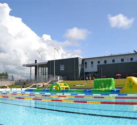 En utomhusbadplats med en stor swimmingpool omgiven av gröna gräsmattor och solstolar. Människor njuter av fritidsaktiviteter i solen på Djupadalsbadet Bad och Camping.