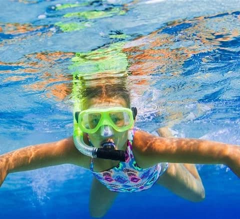 En person med dykutrustning och snorkel glider genom vattnet och har roligt. I bakgrunden syns en campingplats vid vattnet med människor som njuter av semestern vid Djupadalsbadet Bad och Camping.