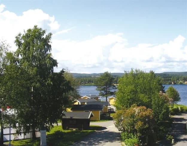 Alt-text: "Vy över vattendrag vid Bergeforsparkens Camping & Stugby, omgiven av grönskande strandkanter och molntäckt himmel, med små stugor intill vattnet."