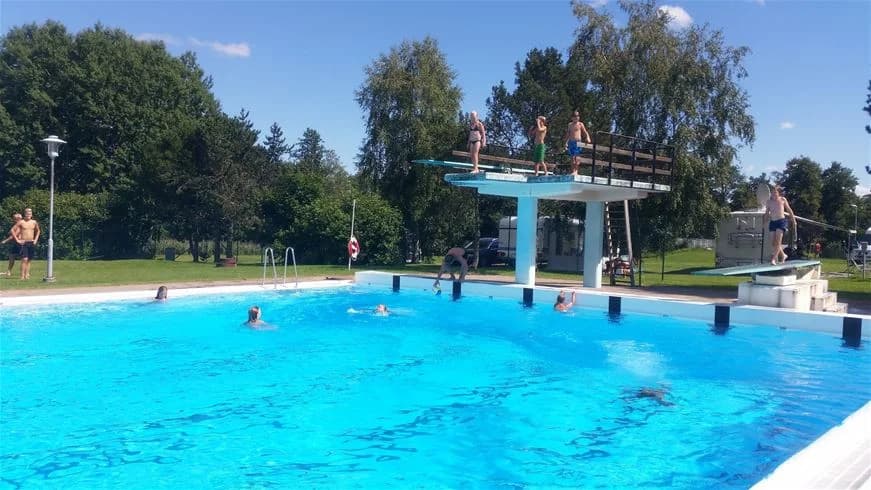 En person dyker ner i en utomhuspool på Nossebro bad & camping, omgiven av grönskande parkmiljö.