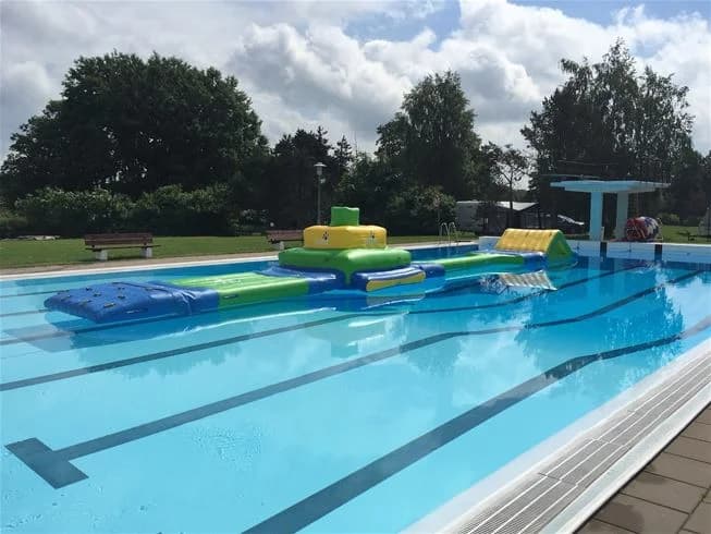 Bild av Nossebro Bad & Camping: En utomhuspool omgiven av grönska och semesterfirare som njuter av fritidsaktiviteter. Solstolar står under skuggande parasoller, och atmosfären är avslappnad och lekfull, perfekt för en semestervistelse.