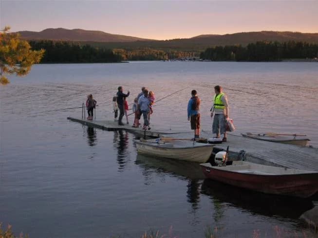 En lugn sjö med flera båtar vid stranden på Sollerö camping, omgiven av grönskande natur och spegelblankt vatten.