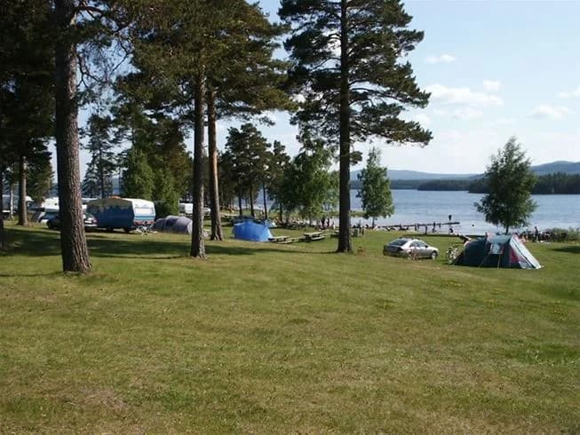 En campingplats vid en sjö med husvagnar och tält uppställda nära vattnet, omgiven av en frodig grönska.