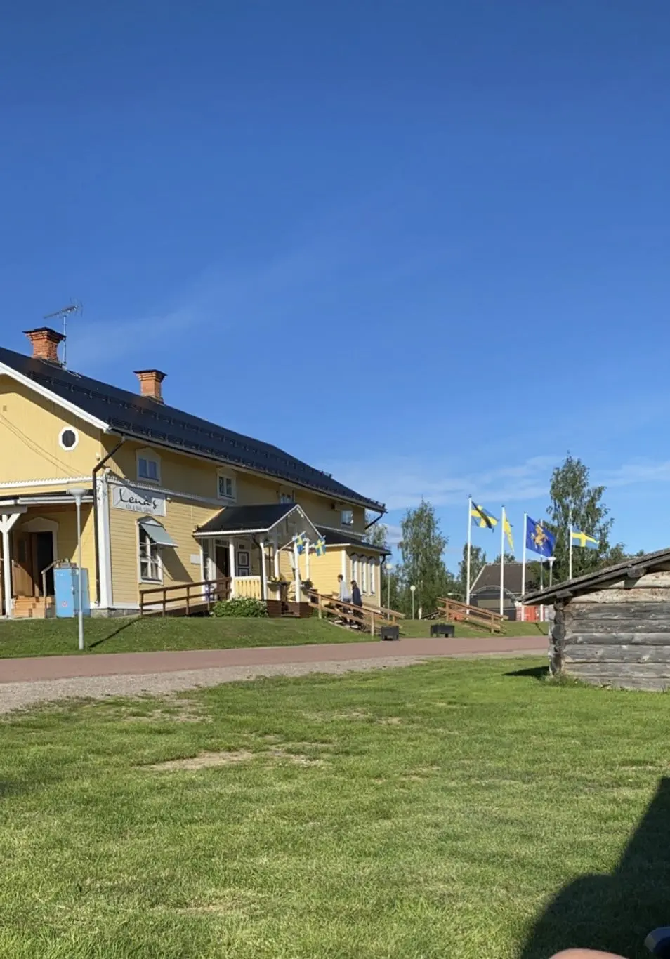 En grönskande campingplats i Särna med plana gräsytor och träd som ger skugga. I bakgrunden syns en byggnad som fungerar som värdshus.