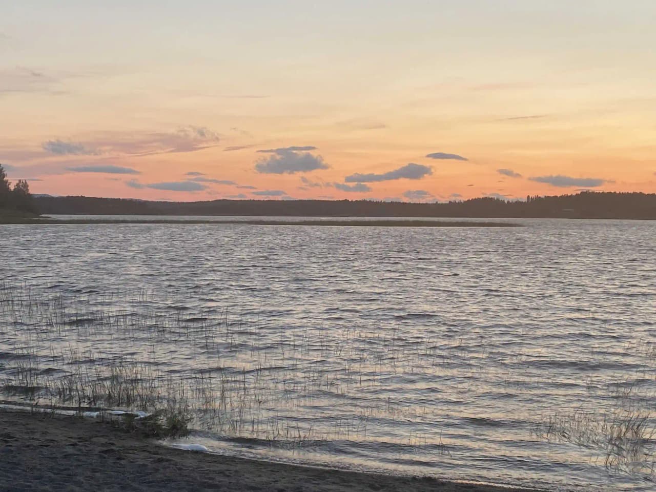 Bild av Särna Camping med en vacker solnedgång i bakgrunden. Himlen är färgad i mjuka nyanser av orange och rosa och speglas i det stilla vattnet. Naturlandskapet runt omkring ger en fridfull känsla.