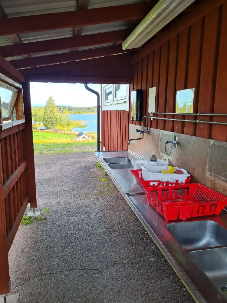 En träbyggnad med mörkare träpanel och ett plåttak omgiven av ett grönskande område på Särna Camping.