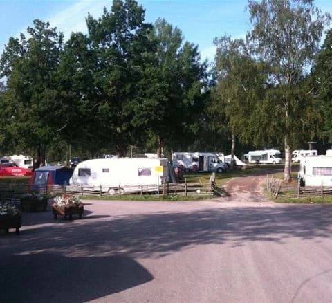 En campingplats med flera uppställda husbilar och husvagnar i en parkeringsliknande miljö. Olika typer av fordon syns i området, inklusive skåpbilar och SUV:ar, omgivna av grönska.