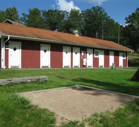 En grön campingplats med rymliga gräsmattor och träd som ger skugga. En välskött uppfart leder genom området, omgiven av trädgårdsliknande landskap och parkbyggnader.