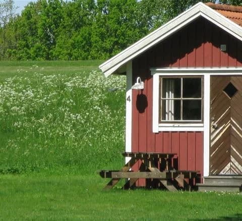 Bild av Östanå camping som visar en stor gräsmatta med ett litet skjul och en stuga i bakgrunden. Olika trädgårdsbyggnader syns runt om på den landsbygdspräglade platsen med gröna marker.