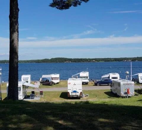 En solig dag vid Örnäs camping med utsikt över en sjö där flera små båtar ligger förtöjda vid stranden. Den natursköna omgivningen visar kust och havslandskap i bakgrunden.