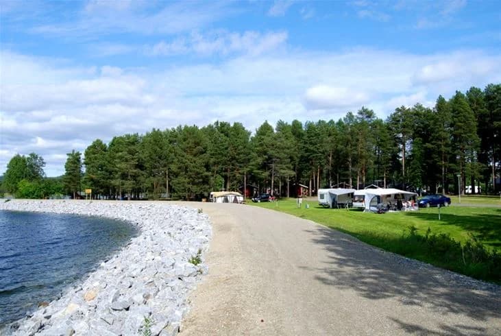 Bild av Storumans Camping vid en naturskön kustlinje. Här syns en strand med småsten intill en klar sjö, omgiven av träd. I bakgrunden breder sig en park ut.