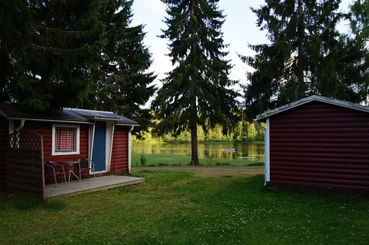 En rofylld campingplats med grönskande träd och välskött gräsmatta omger en mysig stuga i trä. Platsen har en lantlig känsla med trädgårdsbyggnader och landskapsarkitektur som inbjuder till avkoppling och naturupplevelser.