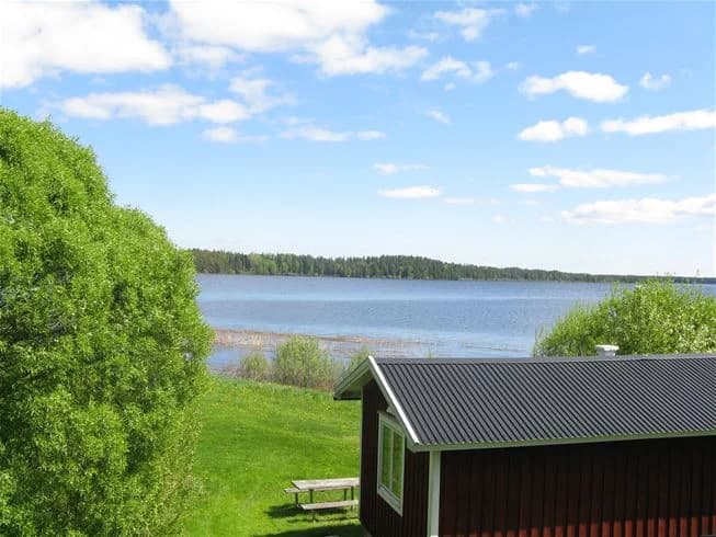 En naturskön bild av Falkudden Camping med stugor vid en sjö. Gröna gräsmattor sträcker sig ner mot strandkanten där lugnt vatten glittrar. Perfekt för friluftsliv och avkoppling.