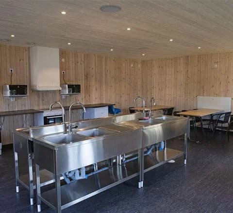 Alt-text: Interiör av ett kök med träpaneler på väggar och tak på Ivö Camping, det finns en taklampa och en köksbänk med silverfärgade inslag.