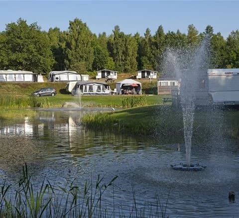 En campingplats med husvagnar och fritidsfordon omgivet av grönskande våtmarker och en liten damm.