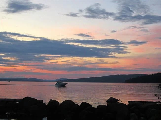 En panoramavy över en stilla sjö vid skymning med glödande solnedgångshimmel. En båt flyter på vattnet, omgiven av skyar och horisontens mjuka linjer.
