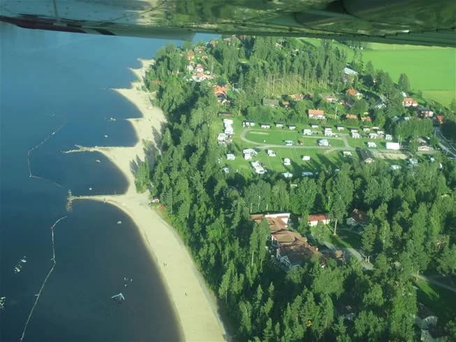 Vy över Orbadens camping med en strandlinje mot vattnet, båtar i viken och besökare som njuter av fritidsaktiviteter i parkområdet.