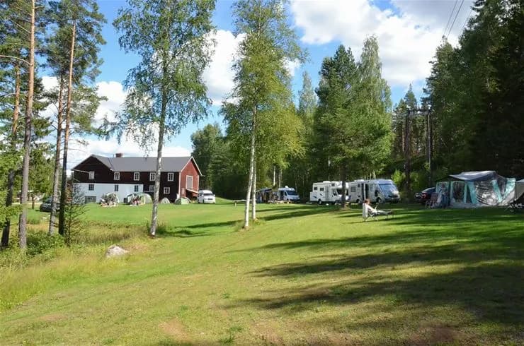 Bild av Wildlife Sweden Camp Ängra med grönt gräs, omgivande träd och växter. Ett litet hus syns i landskapet, vilket ger en känsla av landsbygd och naturlig idyll.