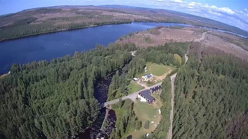 Flygfoto av Wildlife Sweden Camp Ängra, med en vidsträckt skogsmiljö som omger ett öppet område. En flod slingrar sig genom landskapet, vilket ger en blandning av vatten och grönska.
