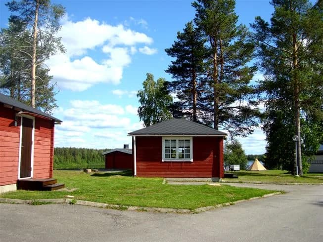 Bild av Pajala Camping Route 99 som visar ett naturskönt landskap med en liten stuga omgiven av gräs. En välskött gräsmatta breder ut sig runt stugan, vilket ger en inbjudande och lantlig atmosfär.