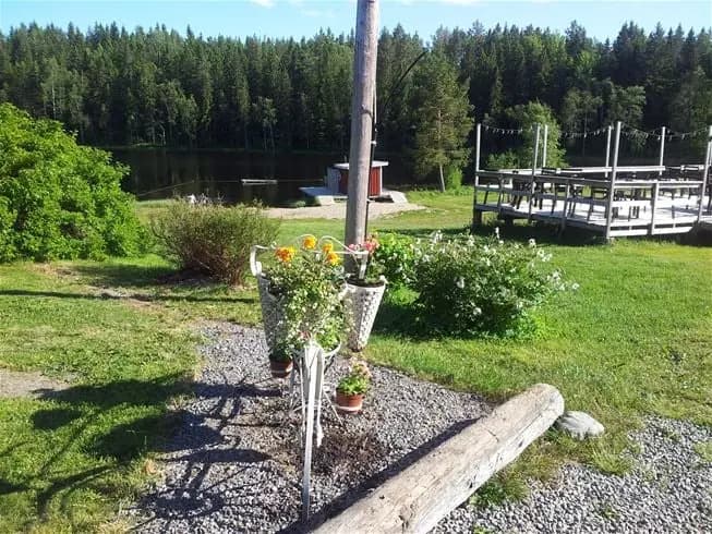 En grönskande trädgård med utemöbler placerade på en välklippt gräsmatta. I bakgrunden syns ytterligare en sittplats med ett picknickbord och bänkar, inramade av lummig växtlighet. En fridfull miljö perfekt för avkoppling och sociala aktiviteter.
