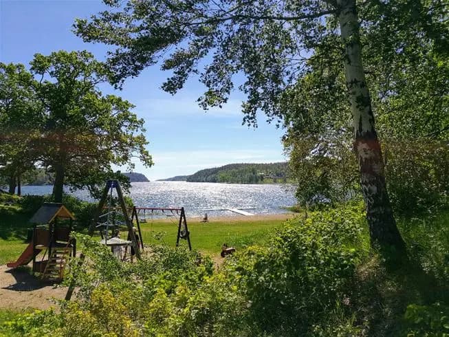 Somrig campingplats vid en sjö, omgiven av gräs och växtlighet med parkbänkar i skuggan vid stranden.