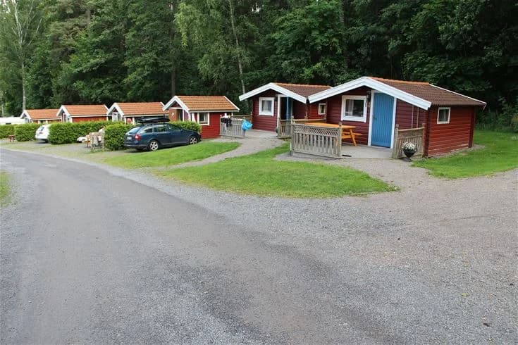 Bild av Malmköpings Bad & Camping: En stuga omgiven av en grönskande trädgård med välskött gräsmatta. I förgrunden syns en uppfart som leder till stugan. Området är naturskönt och inbjudande, med park- och fritidskänsla.