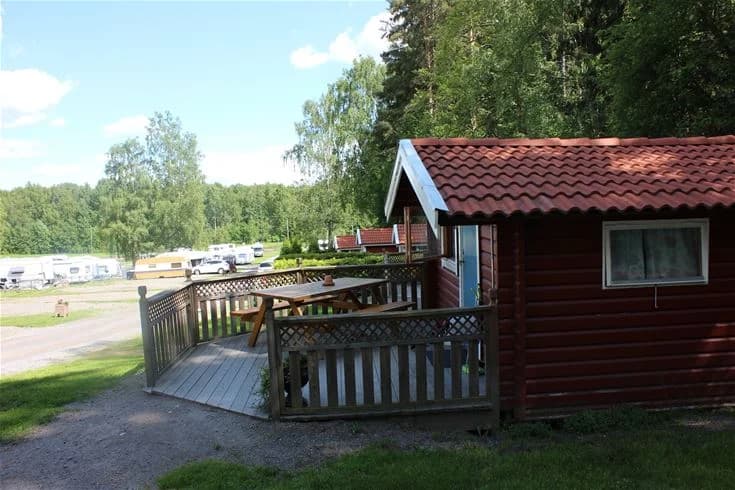 En stuga med en veranda omgiven av grönska på Malmköpings bad & camping. I förgrunden syns en lummig trädgård och en skuggig gårdsplan.