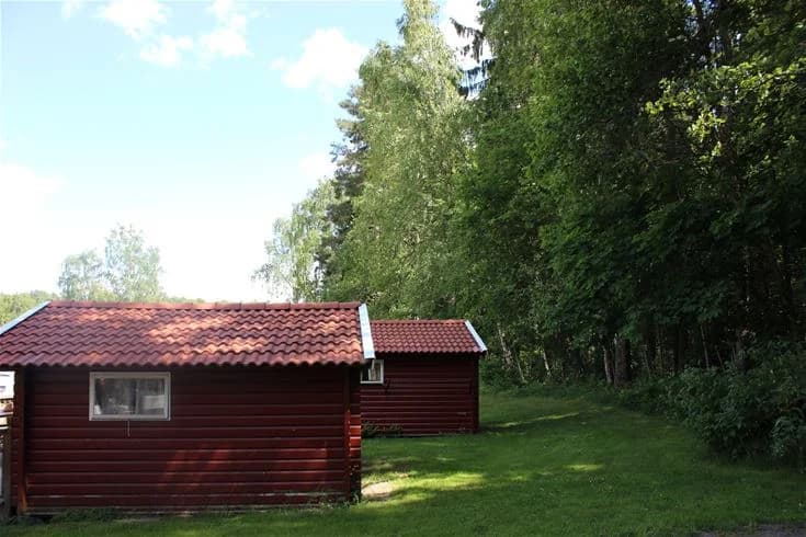 En pittoresk sommarstuga i Malmköpings Bad & Camping omgiven av gräs och träd, med träfasad och tak, vilket ger en inbjudande och lantlig atmosfär.