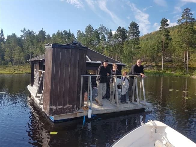 En lantlig stuga vid kanten av en bred vattendrag, omgiven av frodig grönska. En sjö eller flod breder ut sig framför stugan, och en liten båt ligger förtöjd vid stranden. Scenen visar en fridfull och naturnära miljö.
