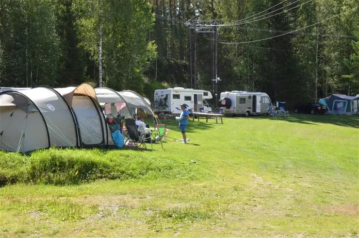 En campingmiljö i skogen med träd och växter runtomkring. Ett tält och en husvagn är synliga, samt en tarp uppspänd som skydd.