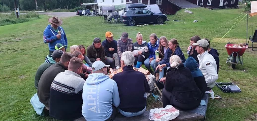 En grupp människor samlade i en avslappnad miljö på Wildlife Sweden Camp Ängra. De sitter på utestolar och vid ett campingbord på en gräsmatta, klädda i jeans och avslappnade kläder och njuter av fritid och gemenskap.